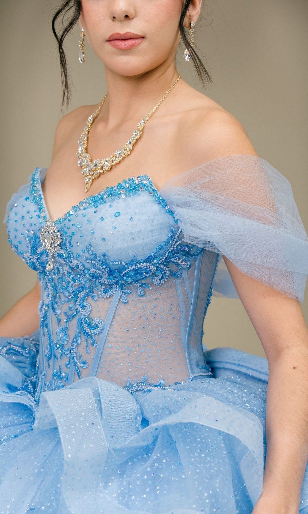 A woman models the Quinceanera Dress 8520JQ By Cinderella Couture—a light blue, beaded off-shoulder gown featuring sheer sleeves and a corset bodice for elegant structure—styled with a statement necklace and photographed from the shoulders to the waist.